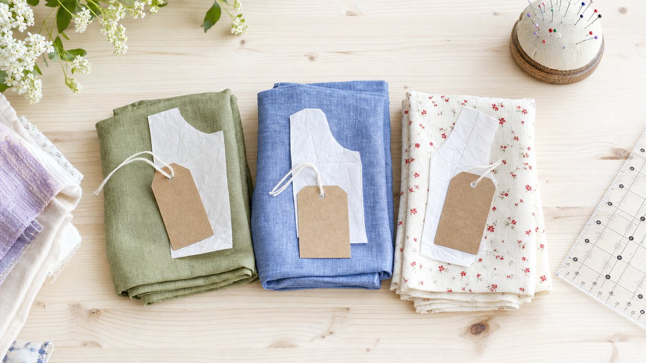 Folded fabric lengths in olive linen, cornflower chambray and a cream floral lawn sorted into small piles on a sewing table