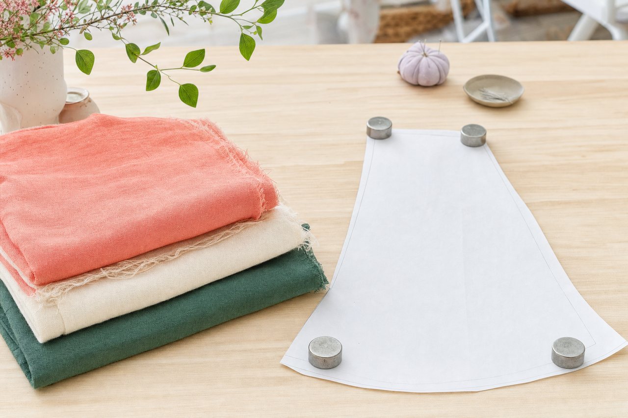 A stack of folded cotton remnants in coral, cream and forest green beside a skirt pattern piece and pattern weights