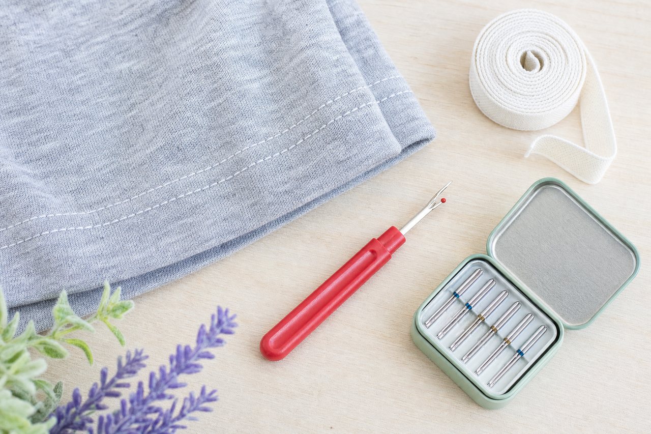 A heather gray jersey hem with twin-needle stitching laid flat beside a strip of knit stay tape and a seam ripper
