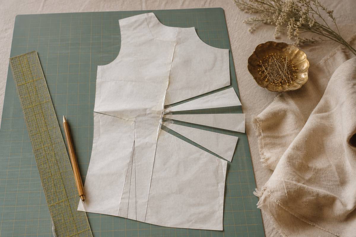 A tissue sewing pattern bodice piece slashed and spread for a full bust adjustment, with ruler, pencil, and pins on a cutting mat
