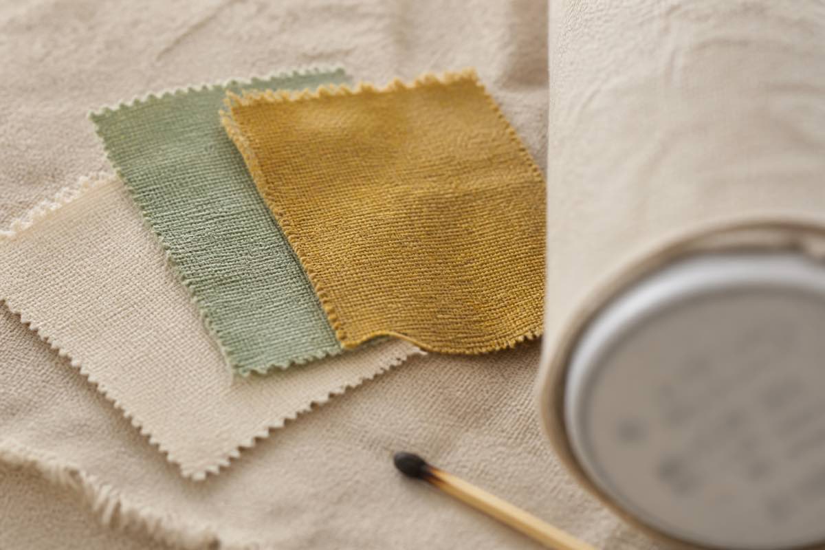 A close-up of a fabric bolt end label showing fiber content percentages and care symbols, beside swatches of unlabeled fabric and a lit match for a burn test