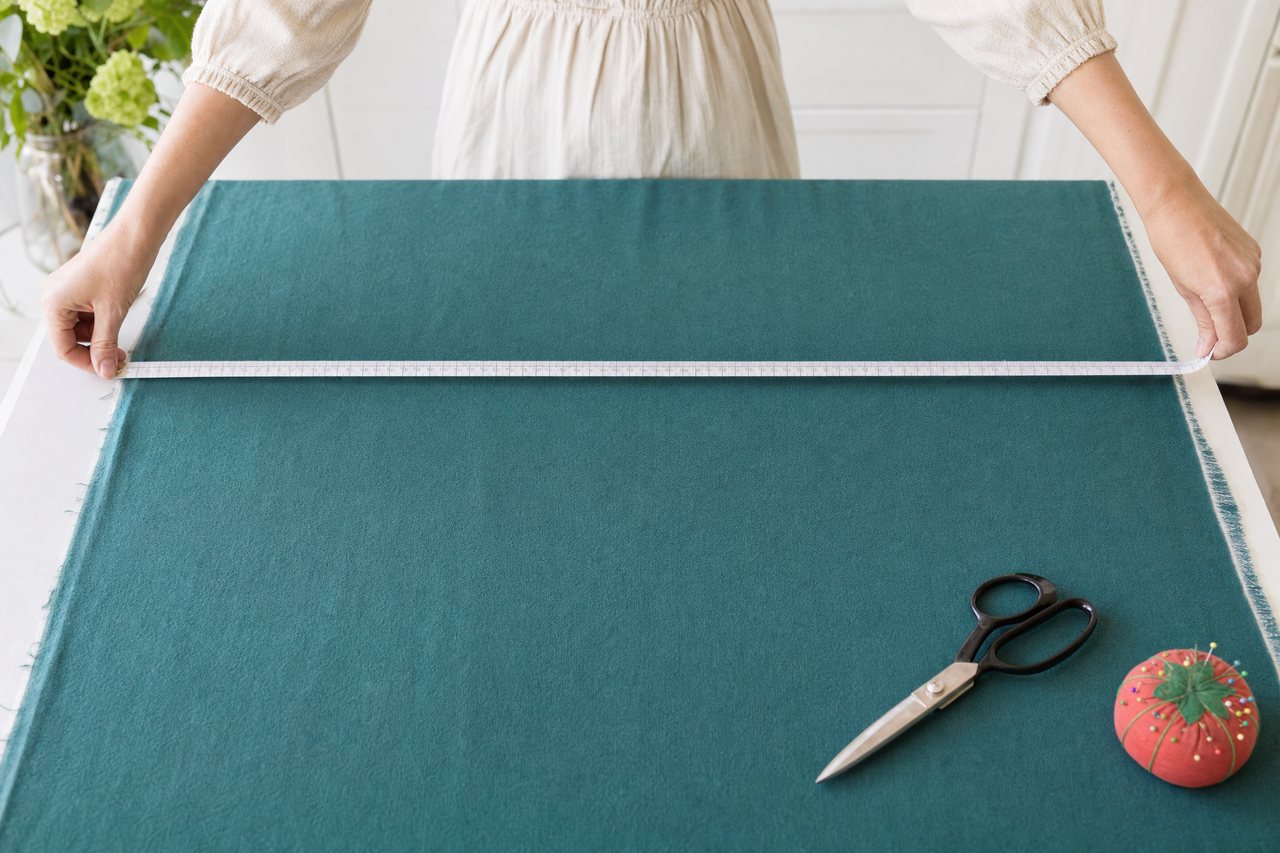 Hands measuring the width of a teal wool crepe with a centimeter tape measure across a cutting table
