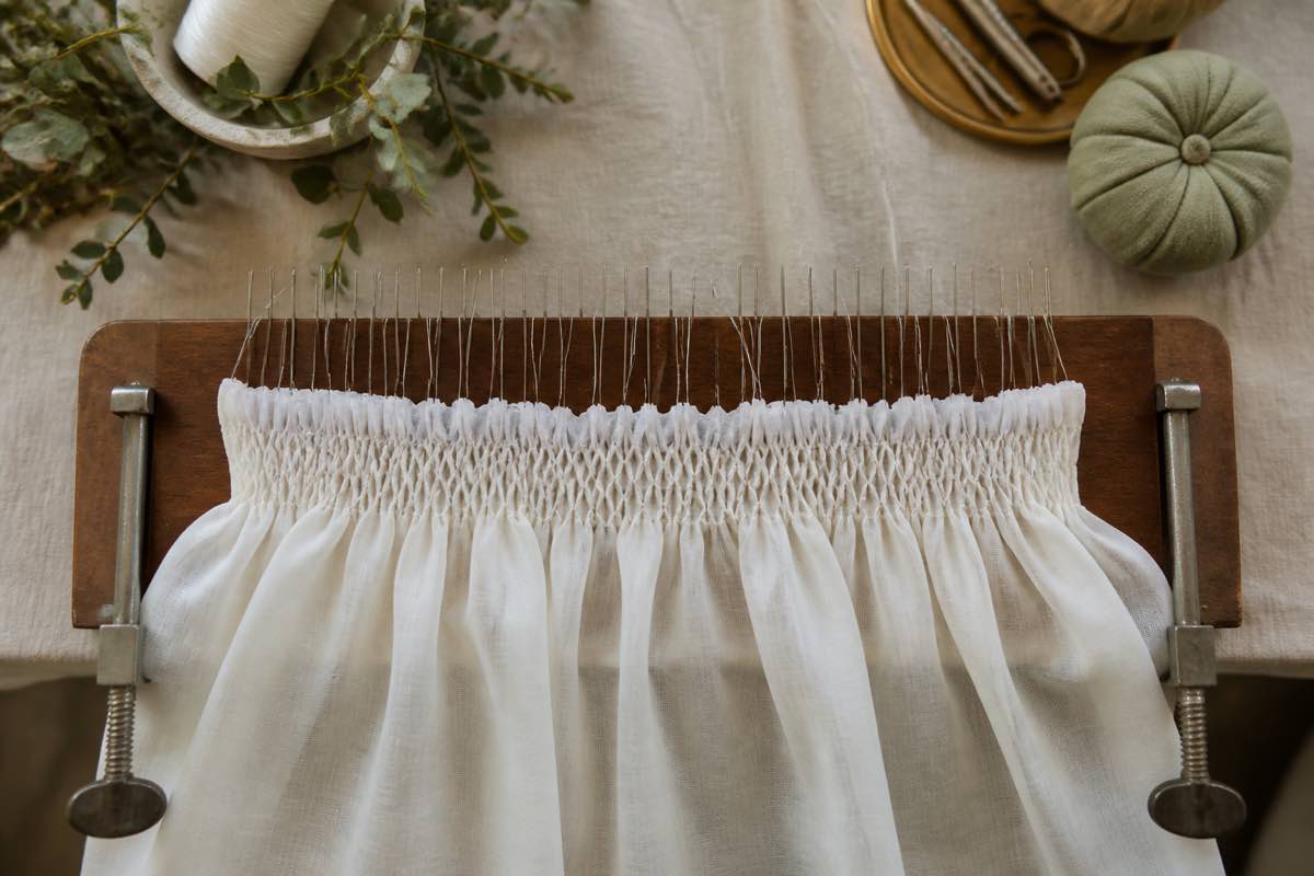 Read smocking pleater clamped to a table with threaded needles ready to pleat batiste fabric