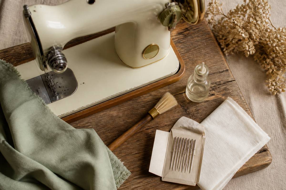 Vintage cream sewing machine on a wooden workbench with a soft brush, small bottle of sewing machine oil, pack of needles, and folded lint-free cloth beside it in warm natural light