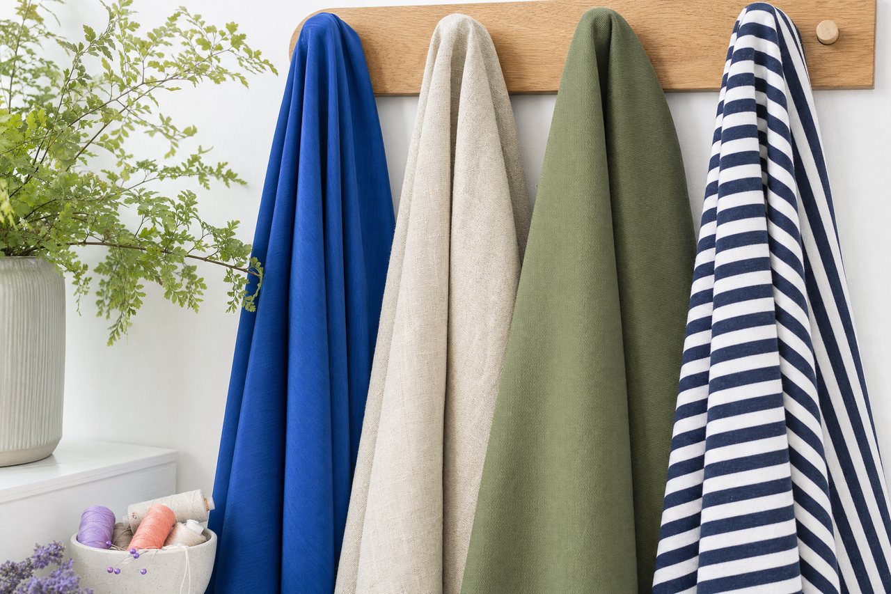 Four fabrics hanging in a row from a wooden rail: rayon, linen, cotton twill and a striped jersey, each falling differently