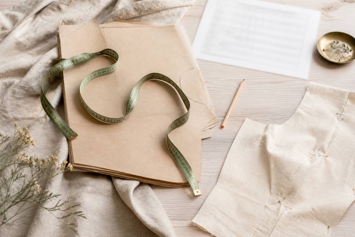 Flexible tape measure draped over a folded paper sewing pattern and size chart on a pale wood table, with a marking pencil and pinned muslin