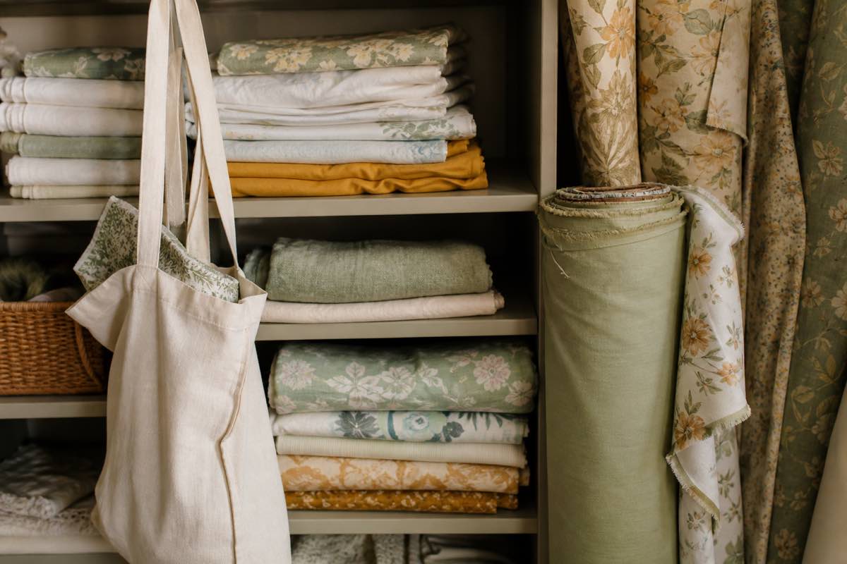 A thrift store shelf of folded bed sheets and fabric remnants with a tote bag, beside vintage sheets and a deadstock fabric bolt
